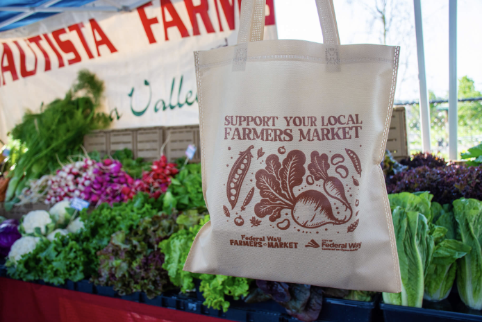 A tote bag advertising the farmers market in front of a table of green vegetables.